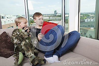 Boy dressed in dinosaur costume sitting with father reading story book on sofa bed at home Stock Photo