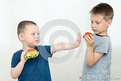 The boy eats a hamburger and refuses fruit offered by a friend. 2 boys on a light background. The concept is to lead a healthy Stock Photo