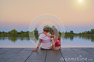 Boy with flat cap and little girl are sitting on pier. Little girl is a hug her older brother. Love, friendship and Stock Photo