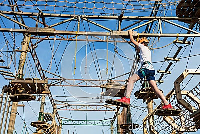 Boy in forest adventure park. Kid in orange helmet and white t shirt climbs on high rope trail. Climbing outdoor Stock Photo