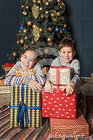 Boy and girl, brother with sister, siblings are sitting near the Christmas tree smiling happily hugging gifts boxes Stock Photo