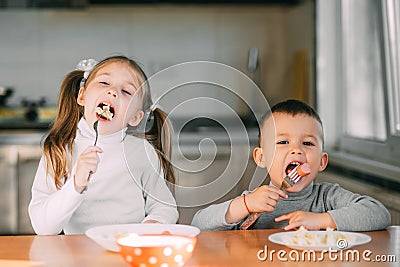 Boy and girl children in the kitchen eating sausages with pasta is very fun and friendly Stock Photo