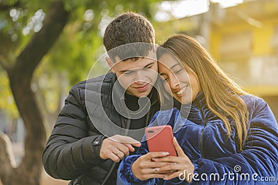 Boy and girl in love sitting on a bench in a public park looking at a mobile phone Stock Photo