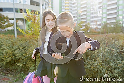 Boy and girl pupils in primary school with digital tablet. Outdoor background, children with school bags, look at the tablet Stock Photo