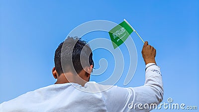 Boy holding Saudi Arabia flag against clear blue sky. Man hand waving Saudi Arabian flag view from back, copy space Stock Photo