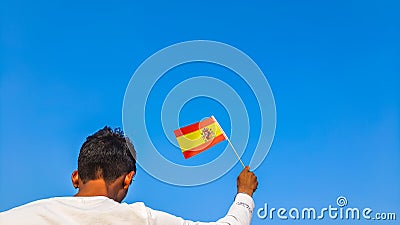 Boy holding Spain flag against clear blue sky. Man hand waving Spanish flag view from back, copy space Stock Photo