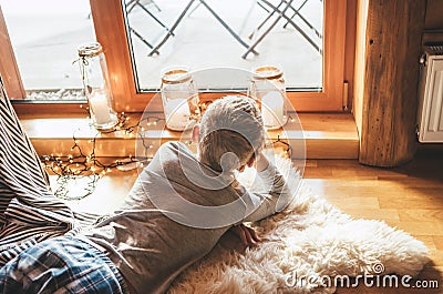 Boy lying on floor on sheepskin and looking in window in cozy home atmosphere. Peaceful moments of cozy home concept image Stock Photo