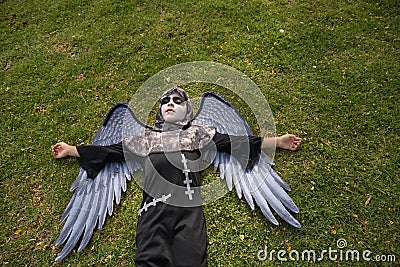 Boy in make up with white face and black eyes, dressed as an angel of death with wings, lying on his back on the grass with a lost Stock Photo