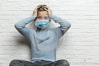 A boy in a medical mask holds his head with his hands. A 9-year-old boy in a blue sweater. Quarantine during the coronavirus Stock Photo