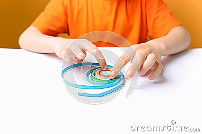 Boy in an orange T shirt sits at a white table and adjusts the twisted thin paper strips with his fingers performs crafts in the Stock Photo
