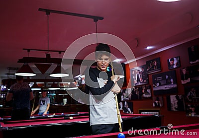 Boy plays billiard or pool in club. Young Kid learns to play snooker. Boy with billiard cue strikes the ball on table. Stock Photo