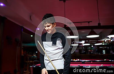 Boy plays billiard or pool in club. Young Kid learns to play snooker. Boy with billiard cue strikes the ball on table. Stock Photo