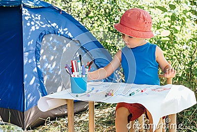 A boy in red Panama sits at a children`s table and draws markers on a white watman in nature near the camping tent Stock Photo