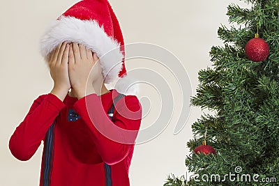 Boy in red Santa cap hidding his face with both hand near christmas tree Stock Photo