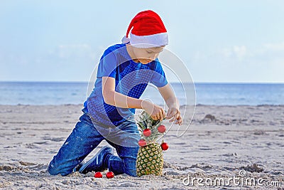 Boy in red Santa hat decorating pineapple as a Christmas tree on a sunny sandy beach by the sea Stock Photo