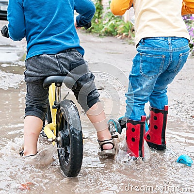 Boy is riding a bike and girl is riding a scooter across deep puddle Stock Photo