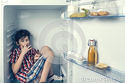 A boy in a shirt and shorts eating a chocolate bar inside a fridge with food Stock Photo