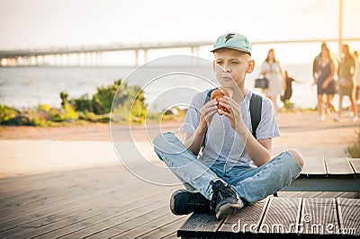 The boy sits on a bench on the waterfront with his legs crossed and eats a croissant. A teenager in a blue cap and T-shirt eats Stock Photo