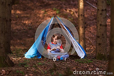 Boy sitting under a play tent in the woods playing alone in solitude enjoying creating a bow play pretending. Stock Photo
