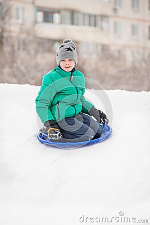 Boy slides down the hill on snow saucer against the background of multi-house. Seasonal concept. Winter day Stock Photo