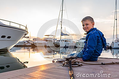 A boy with a smile in a blue jacket sits on a pier with yachts during sunset Stock Photo