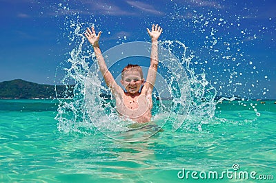 Boy splashing water in sea. Playful child 10 years old surrounded by colorful nature. Bright blue sky and shimmering sea. Stock Photo