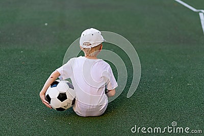 A boy in sports clothes sitting on a green lawn on a football field with a soccer ball back, back view, sports section, training Stock Photo