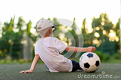 A boy in sports clothes sitting on a green lawn on a football field with a soccer ball back, back view, sports section, training Stock Photo