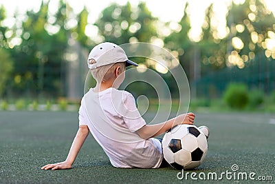 A boy in sports clothes sitting on a green lawn on a football field with a soccer ball back, back view, sports section, training Stock Photo