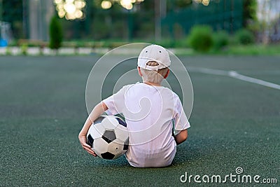 A boy in sports clothes sitting on a green lawn on a football field with a soccer ball back, back view, sports section, training Stock Photo