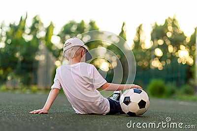 A boy in sports clothes sitting on a green lawn on a football field with a soccer ball back, back view, sports section, training Stock Photo