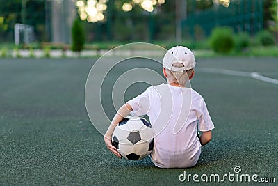 A boy in sports clothes sitting on a green lawn on a football field with a soccer ball back, back view, sports section, training Stock Photo