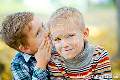 A boy tells a secret to his friend in the ear in an autumn Park against a background of yellow leaves. The boys smile Stock Photo