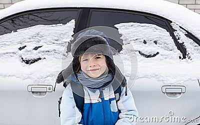 boy in a warm fur hat stands next to a car covered with snow and human faces painted on the windows Stock Photo