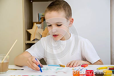 A boy in a white T-shirt sits at a white table and draws with pencils and paints Stock Photo