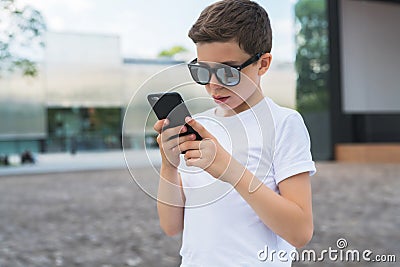 Boy in white T-shirt and sunglasses stands outdoor and uses smartphone. Boy plays computer games on digital gadget Stock Photo