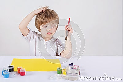 The boy wondered what would draw. He scratches his head. Brush in hand. Paper and paint on the table Stock Photo