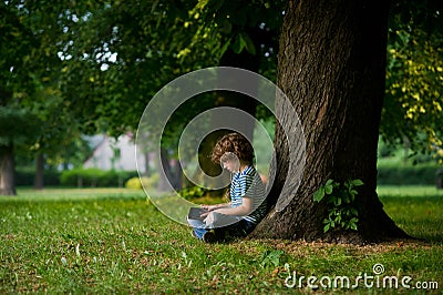 The boy of 8-9 years sits under a big tree, having inclined over the tablet. Stock Photo