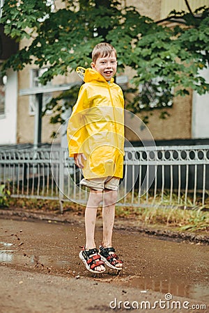 A boy in a yellow raincoat walks outside in the rain. A child jumps on the narrows. jump through muddy puddles. Stock Photo