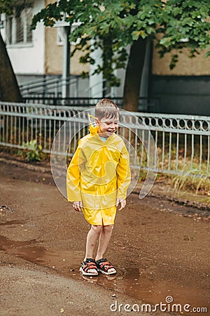 A boy in a yellow raincoat walks outside in the rain. A child jumps on the narrows. jump through muddy puddles. Stock Photo