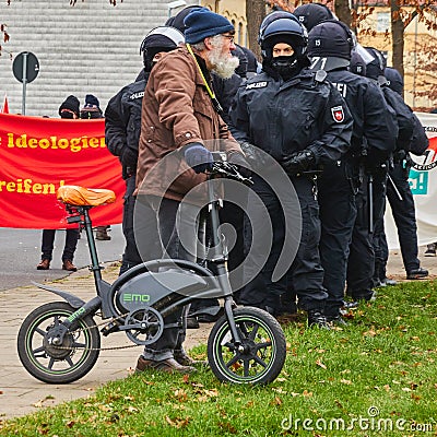 Older man with white beard and blue knitted cap pushes a tiny electric folding bike in front of a group of police officers in Editorial Stock Photo