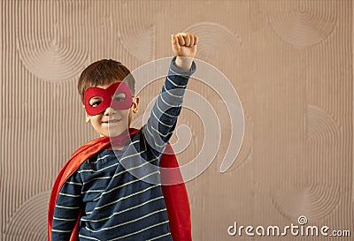 Brave cheerful boy in superhero cape and mask standing with hand on waist on beige background Stock Photo