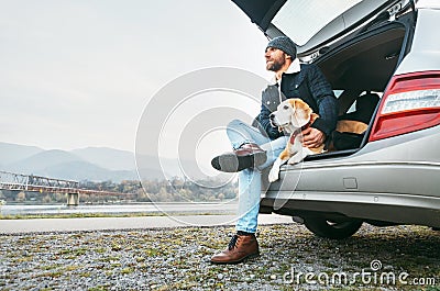 Breaded man in warm clothes siting with beagle in car trunk. Traveling with pet concept image Stock Photo