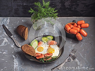 Breakfast of fried eggs and sausages on a black ceramic plate. In a small plate a portion of sausages for the next preparation Stock Photo