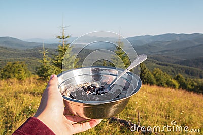 Breakfast in the hike, a bowl of porridge and berries in the female hand on the background of the morning mountain landscape Stock Photo