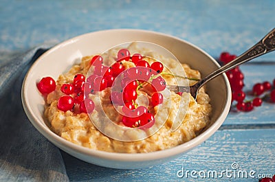 Breakfast of milk oatmeal with red currants in a deep white plate on a wooden tray, a spoon and a denim napkin, as well as sprigs Stock Photo