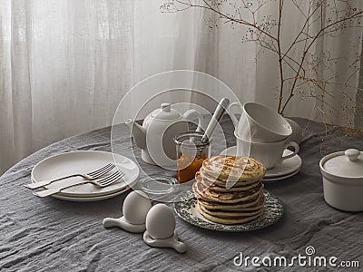 Breakfast served on the table - pancakes, apricot jam, boiled eggs in ceramic egg stands, breakfast dishes on the table with a Stock Photo