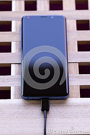 Brecht, Belgium - april 1 2020: A portrait of a black and blue smartphone lying on a wooden box with a USB cable connected to its Stock Photo