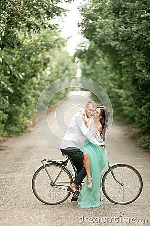 Bride and groom sit on bicycle on forest road, embrace and kiss. Stock Photo