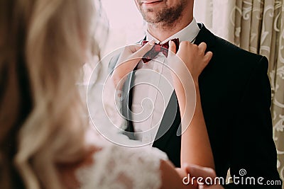 Bride fixes red bow tie on groom`s neck while they stand near the window Stock Photo